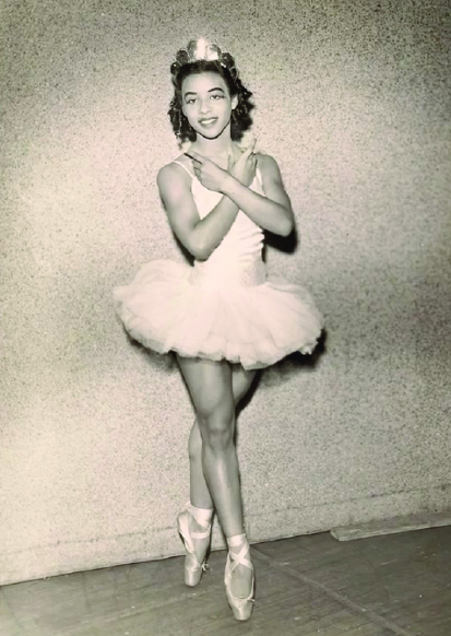 An early image of Delores Brown. She wears a crown in her curly brown hair and dons a white short tutu and pointe shoes. She stand en pointe with her arms crossed over her chest. 